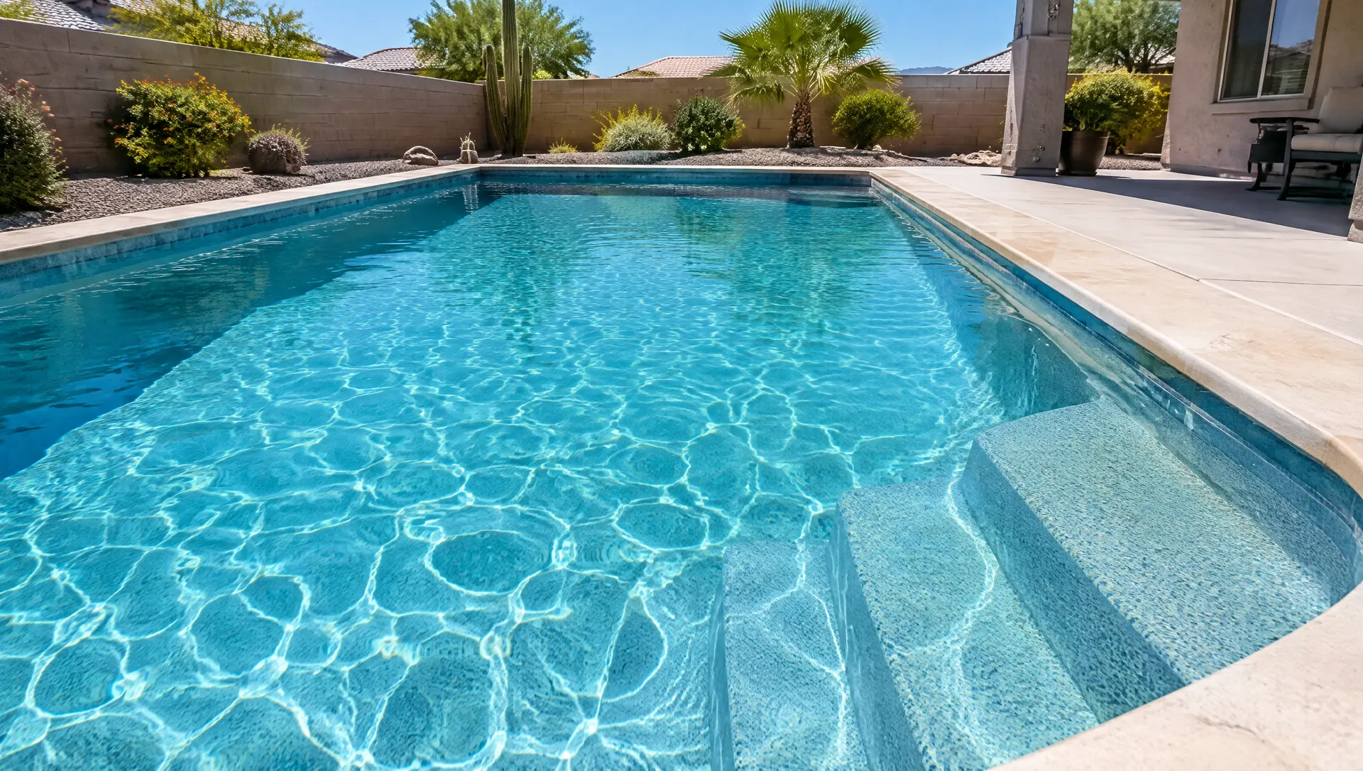 Newly resurfaced residential pool filled with clear water showing a smooth aqua-blue pebble finish on the floor and steps, bright Arizona sun casting caustic light patterns, clean coping edge