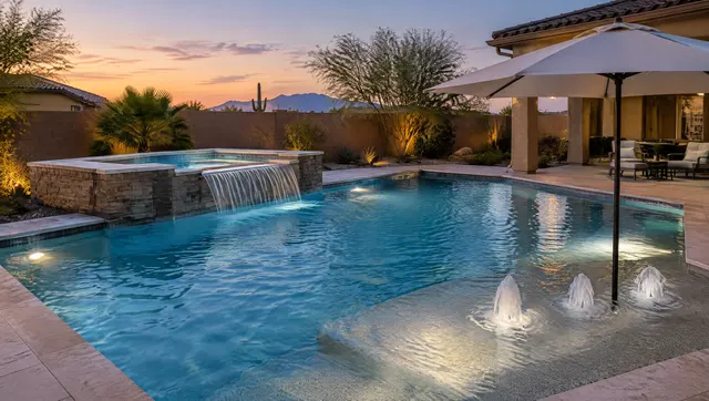 Remodeled backyard pool in Mesa, Arizona with a new raised spa spilling a sheet of water into the pool, a shallow baja shelf with an umbrella, and three bubblers on the shelf, twilight with soft underwater lights